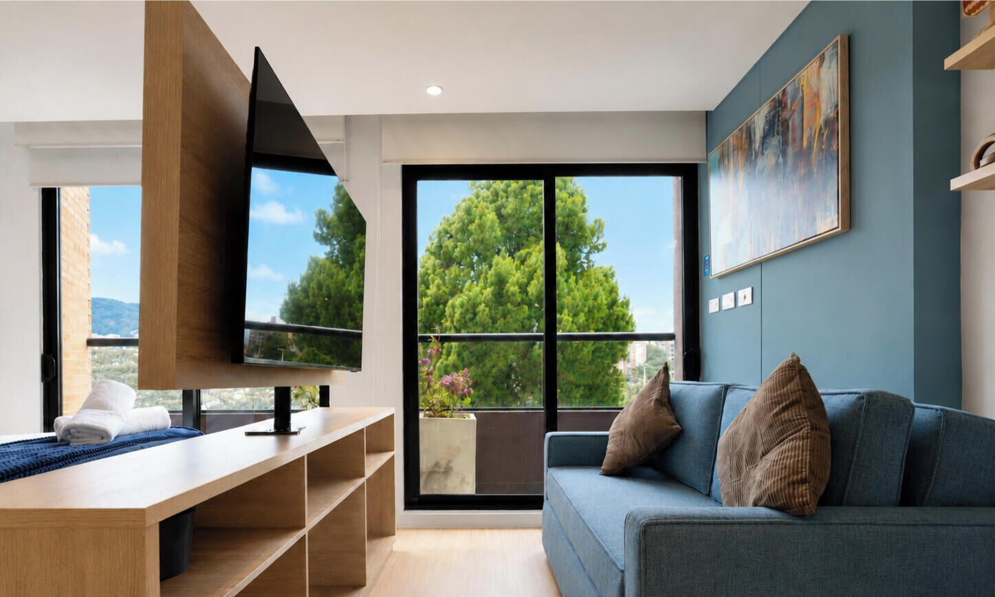 A modern living room with a flat-screen TV, a blue sofa, and a large window offering a view of trees and buildings.