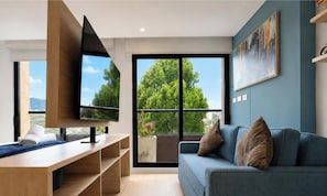 Un soggiorno moderno con una TV a schermo piatto, un divano blu e un'ampia finestra con vista su alberi e palazzi.