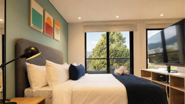 Habitación de hotel con cama, escritorio, lámpara, televisor y ventana con vista a árboles y edificios.