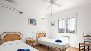 Quarto com duas camas de solteiro, um ventilador de teto, janela com grades e uma cadeira.