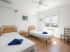 Ein Zimmer mit zwei Einzelbetten, einem Deckenventilator, einem vergitterten Fenster und einem Stuhl.