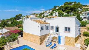 Um edifício branco com base de pedra, piscina e um alpendre com espreguiçadeiras.