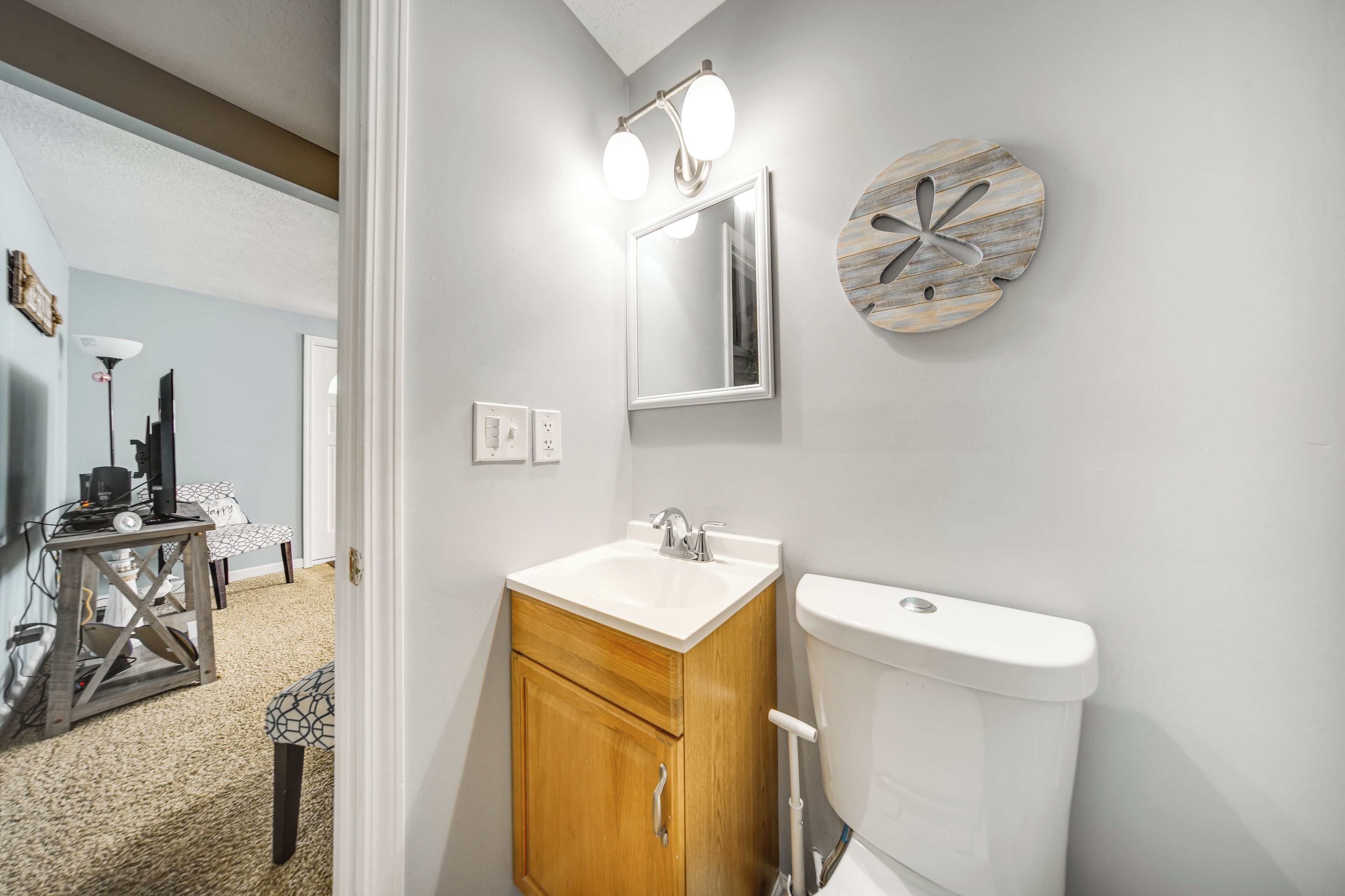 A bathroom with a white sink, wooden cabinet, and a toilet. There is a mirror with lights above it and a decorative wall piece.