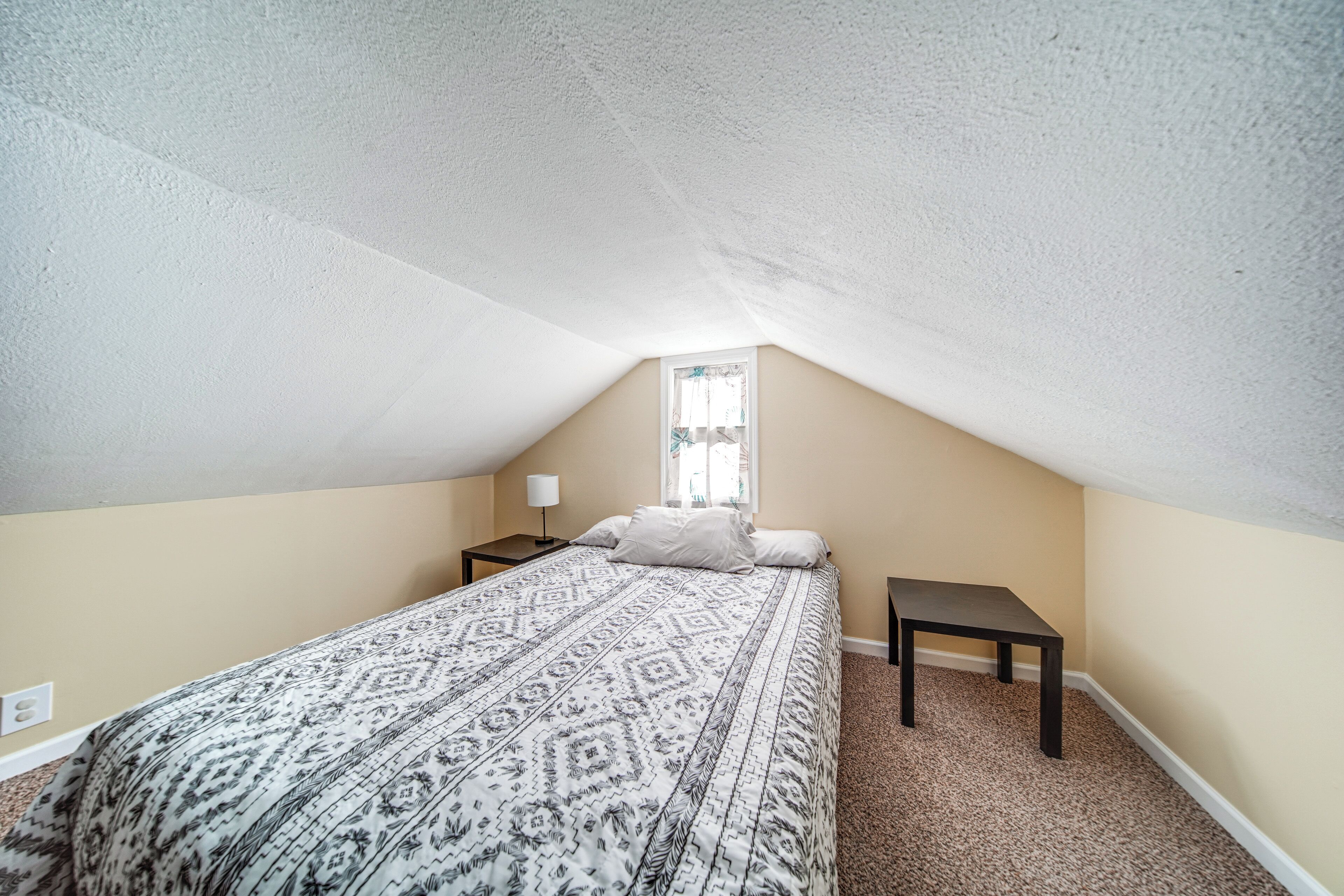 A bedroom with a patterned bedspread, a nightstand, a bedside table, and a window with curtains.
