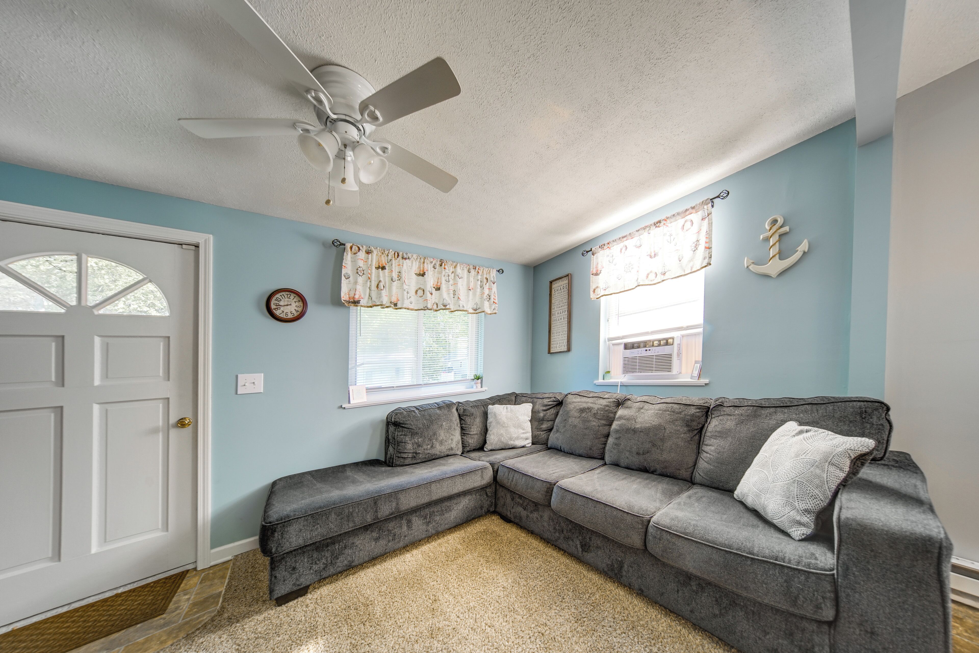 A room with a ceiling fan, a sectional sofa, two windows with curtains, a door, and a small rug.