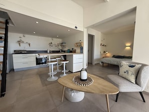 Eine moderne Küche mit weißen Schränken, einer zentralen Kochinsel mit Barhocker und einem Essbereich mit rundem Tisch und einem gemusterten Sofa.