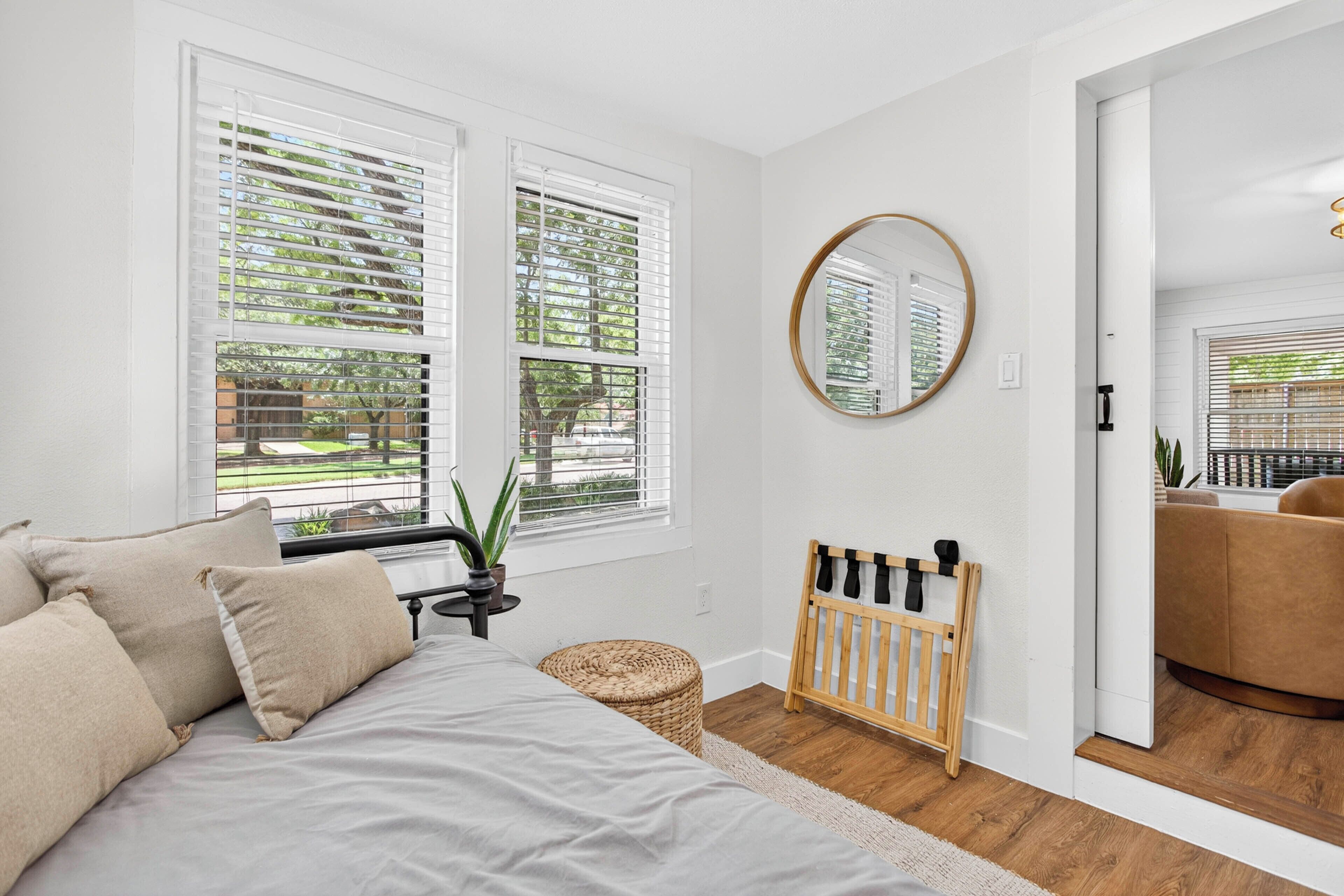 A bedroom with a bed, two windows with blinds, a round mirror, a crib, and a view of the outdoors.