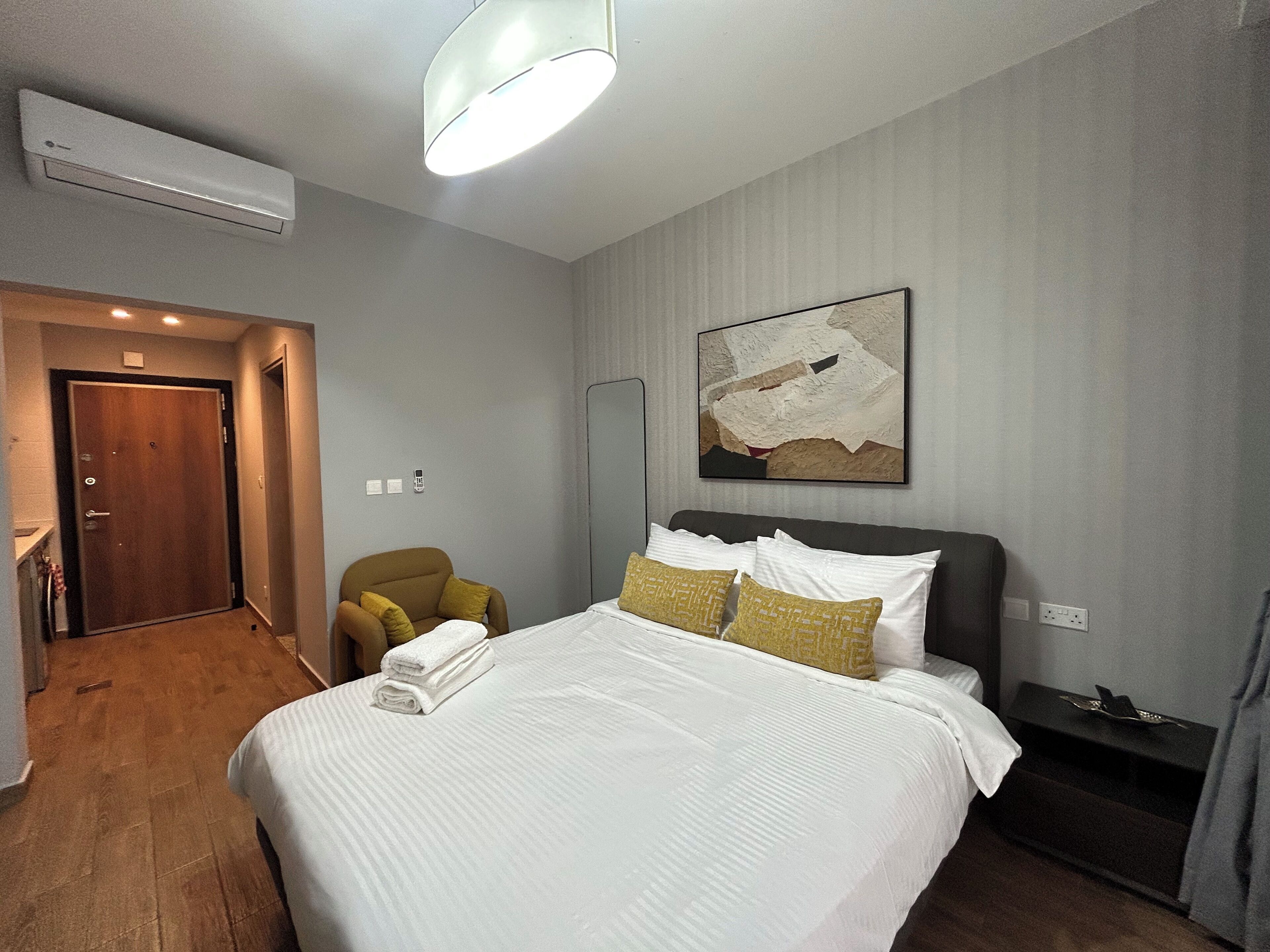A hotel room with a bed, a chair, a bedside table, a wall-mounted artwork, and an air conditioning unit.