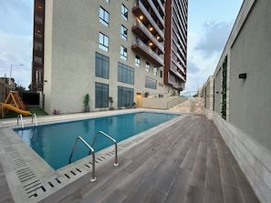 A swimming pool with a metal ladder, surrounded by a building with multiple balconies and large windows. - Lawrounds At HARMONIA RESEIDENCE  (Accra)