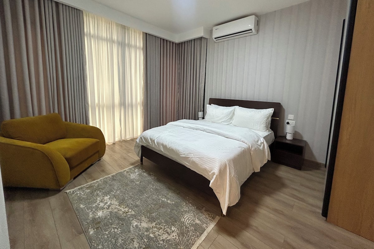 Quarto de hotel com cama, cadeira, mesa de cabeceira, aparelho de ar condicionado e janela com cortinas.