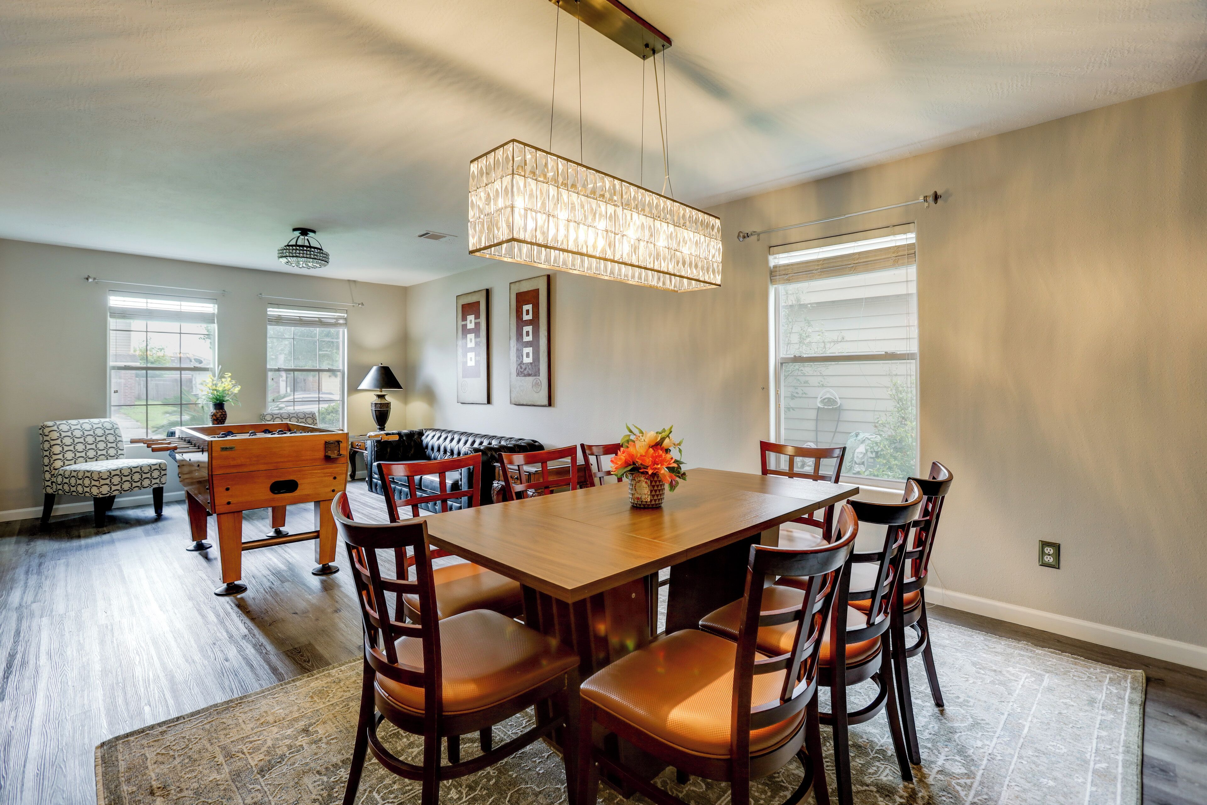 A dining area with a wooden table and chairs, a chandelier, and a window with blinds.