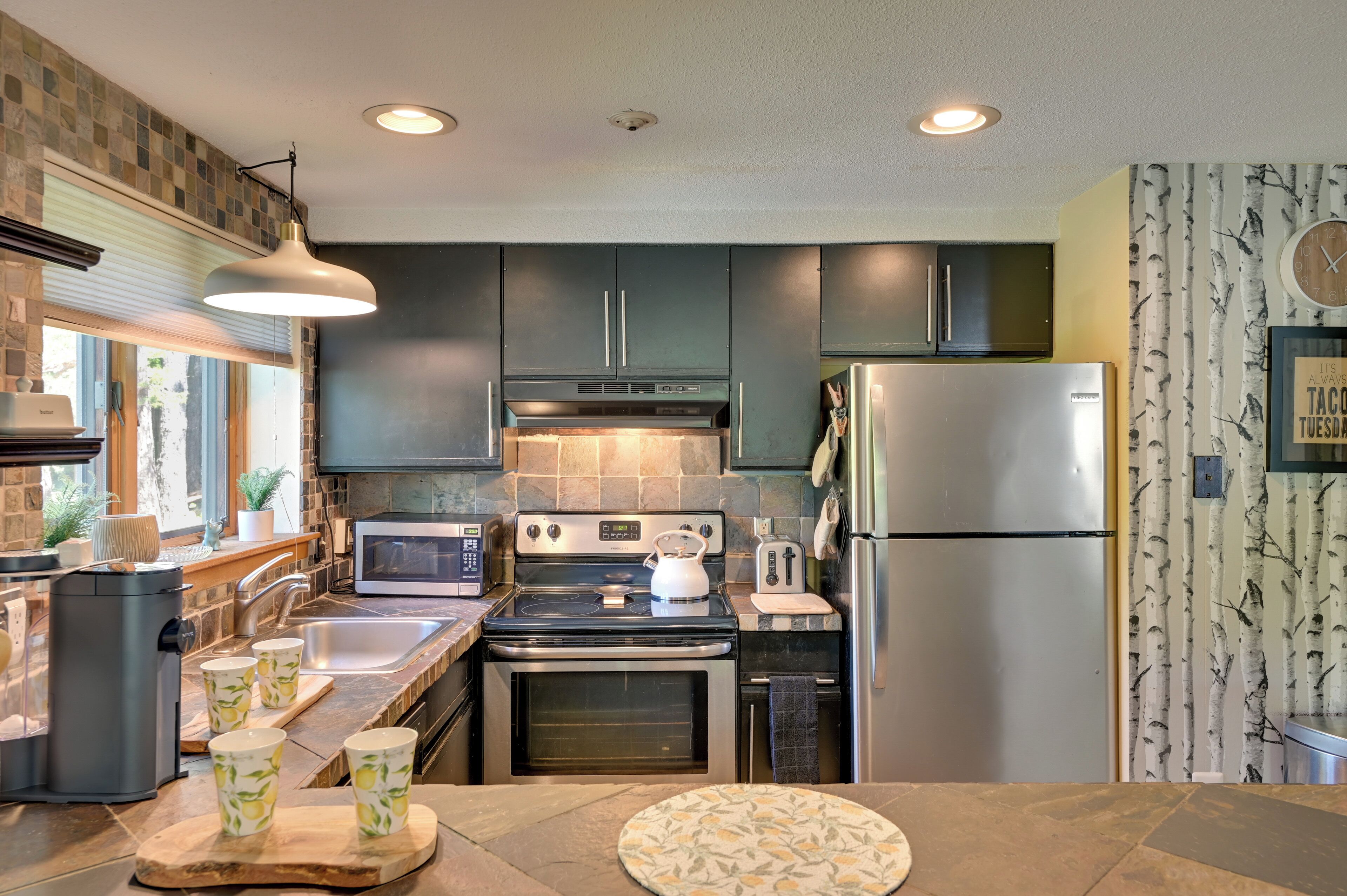 A modern kitchen with dark cabinets, stainless steel appliances, and a large window.