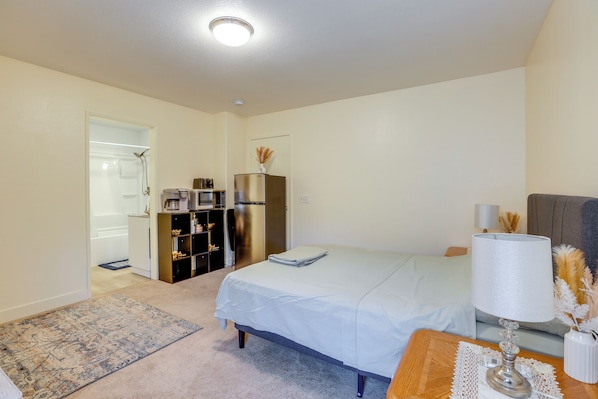 A bedroom with a bed, a nightstand, a lamp, a refrigerator, a microwave, and a bathroom visible in the background.