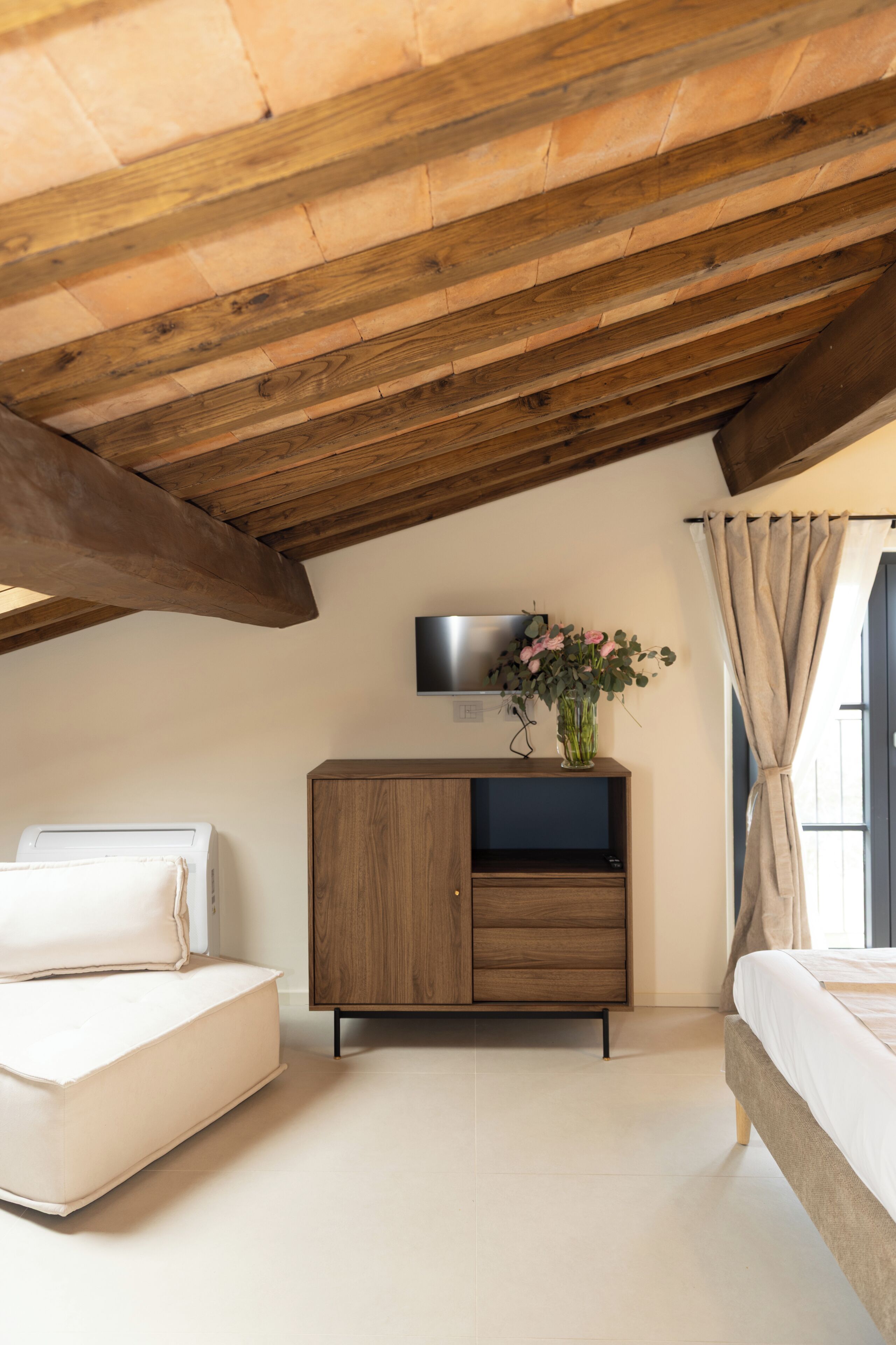Une chambre à coucher avec un plafond en bois, un lit, un canapé, un téléviseur posé sur un meuble et une fenêtre avec des rideaux.