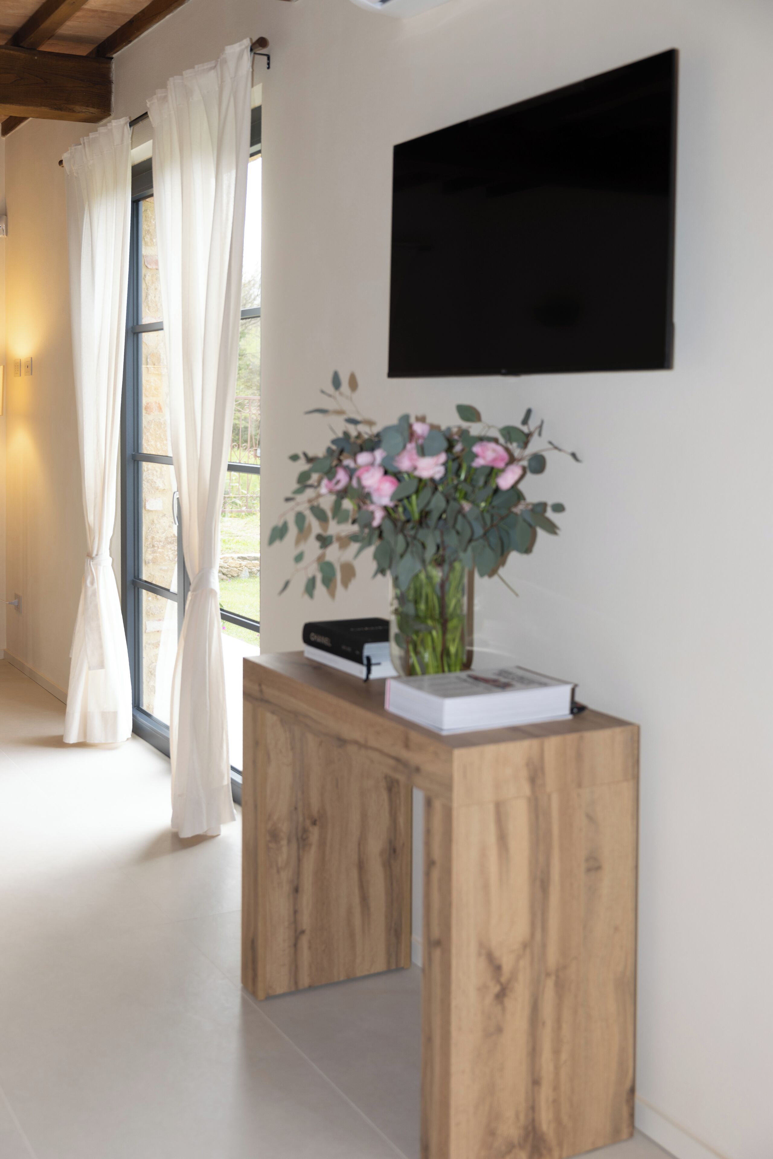 A room with a wooden console table, a vase with flowers, a black framed picture, and a window with white curtains.