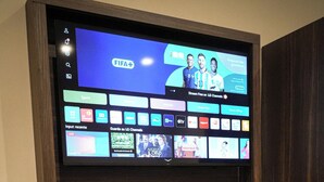 Ein wandmontierter Flachbildfernseher zeigt verschiedene Sender, ein Tisch mit Kaffeemaschine, Gläsern und Fernbedienung.