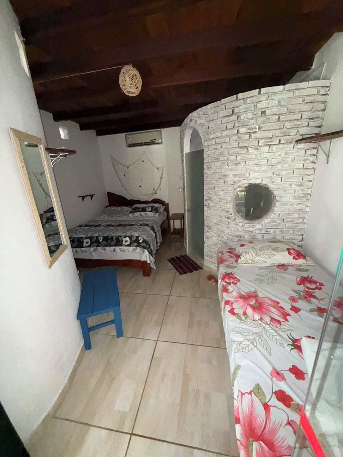 Quarto com duas camas, um banquinho azul, uma parede de tijolos e um espelho.