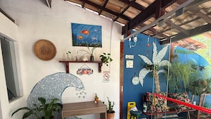 Una habitación con suelo de baldosas, un escritorio de madera, una silla, una planta en maceta, una pared con un cuadro y una puerta de cristal que da al exterior.