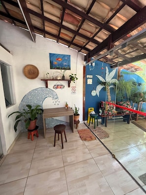 A room with a tiled floor, a wooden desk, a chair, a potted plant, a wall with a painting, and a glass door leading outside. - Arandú Hostel (Itacaré)
