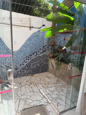 A glass-enclosed shower area with mosaic tile walls and floor, potted plants, and a metal rack. - Arandú Hostel (Itacaré)