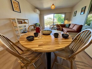 Un área de comedor con una mesa de madera, sillas y cuencos. Se observa una estantería, una jarra roja y un sofá al fondo.