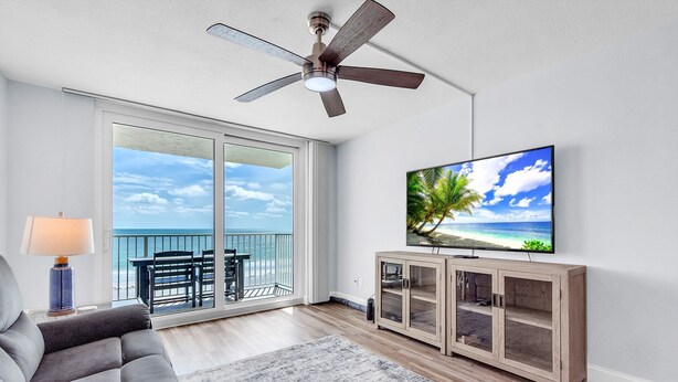 Ein Wohnzimmer mit großem Fernseher, Deckenventilator, Sofa, TV-Stand und einem Balkon mit Meerblick.