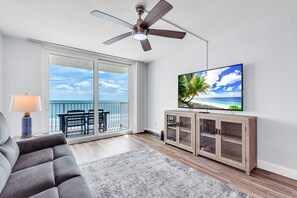 A living room with a large TV, a ceiling fan, a sofa, a TV stand, and a balcony with a view of the ocean. - Beachfront - Heated Pool - Direct Beach Access - Gulf Shores 402 (Indian Shores)