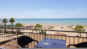 Uitzicht op het strand vanaf een balkon met stoelen en een tafel, palmbomen en parasols op het zand.