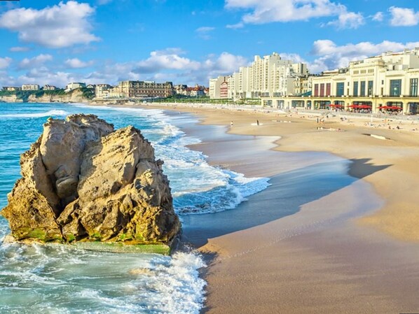 Ein Strand mit einer großen Felsformation, sandigem Ufer und Gebäuden im Hintergrund.