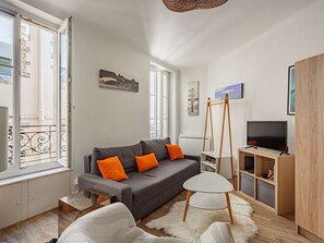 Ein modernes Wohnzimmer mit grauem Sofa, orangefarbenen Kissen, einem weißen Couchtisch, einer TV-Konsole und Kunstwerken an den Wänden.
