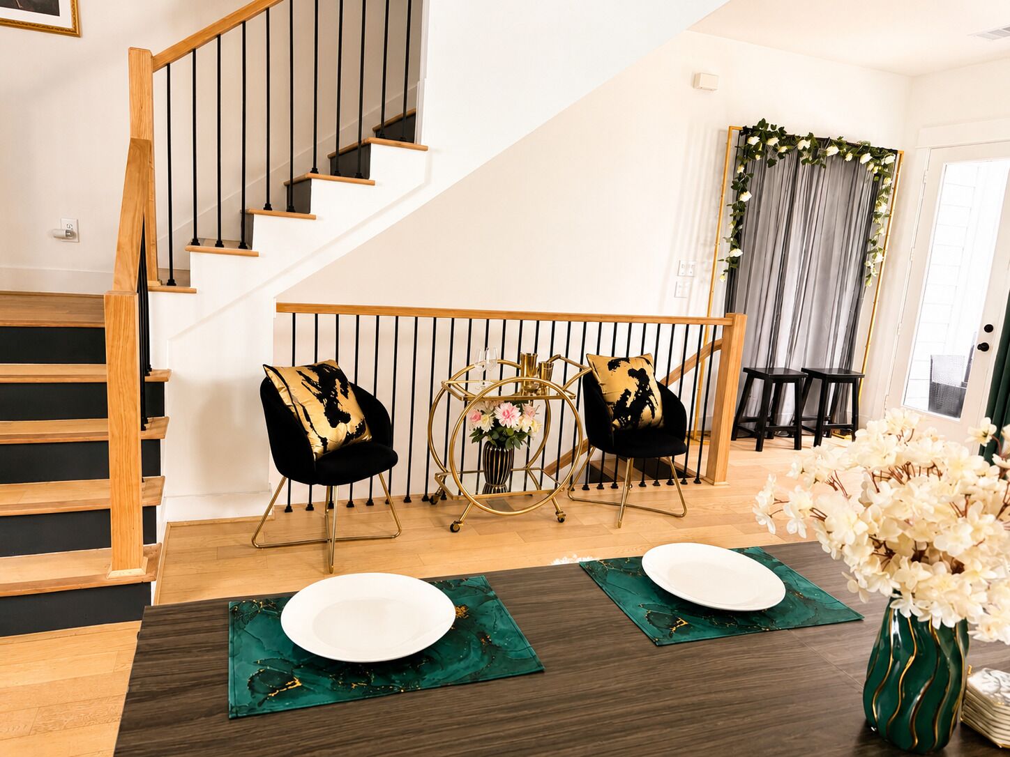 Una escalera con peldaños de madera y barandillas negras, un área de comedor con dos sillas y una mesa, y una cortina decorativa al fondo.