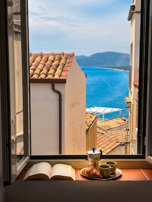 A window view of a coastal town with terracotta roofs, a clear blue sea, and distant mountains.