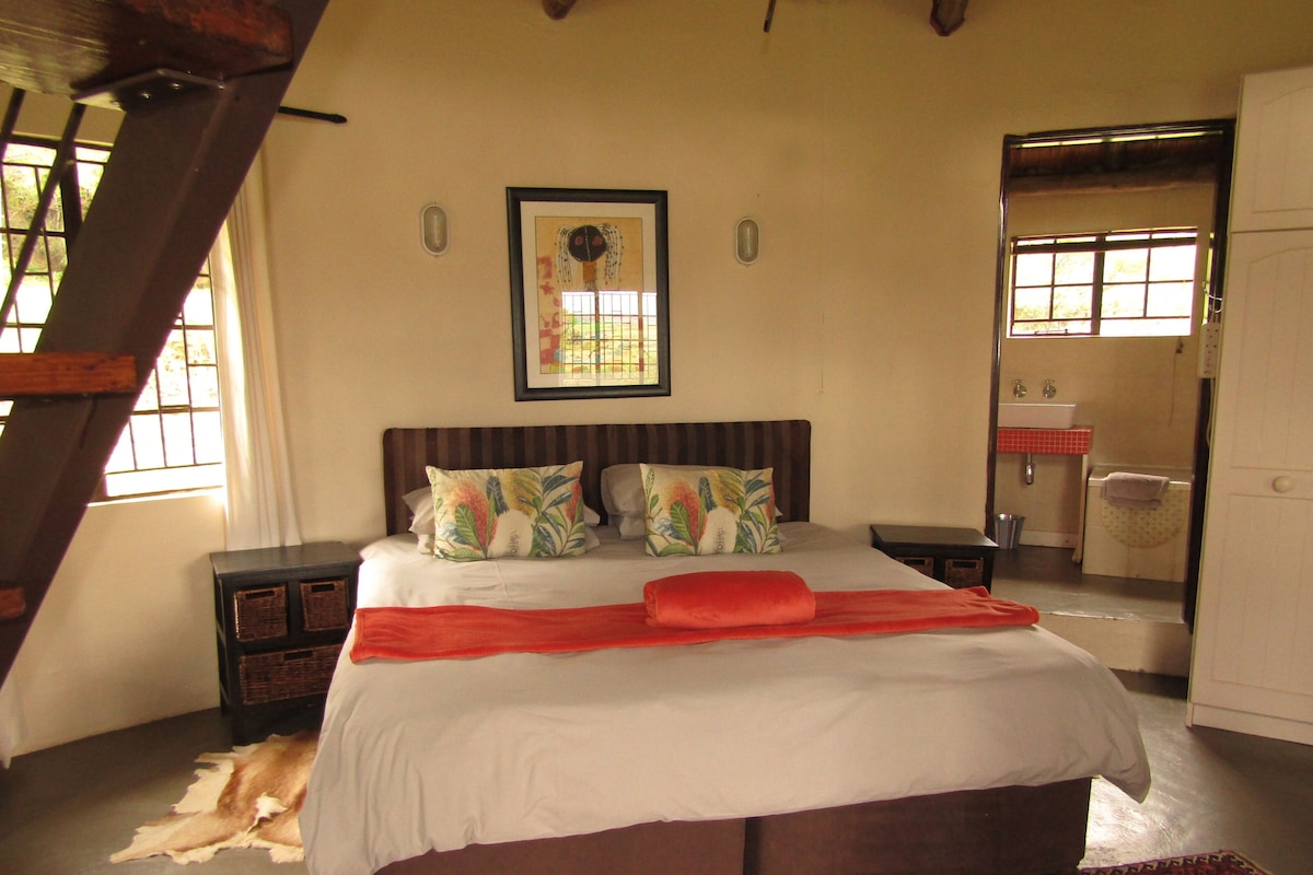 Quarto com uma cama grande, cabeceira de madeira, pintura sobre a cama, janela com cortinas, banheiro com pia e espelho, e um tapete estampado.
