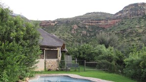 A swimming pool with a stone border, surrounded by grass, with a thatched-roof structure and a fence in the background. - Mafube Mountain Retreat (Fouriesburg)
