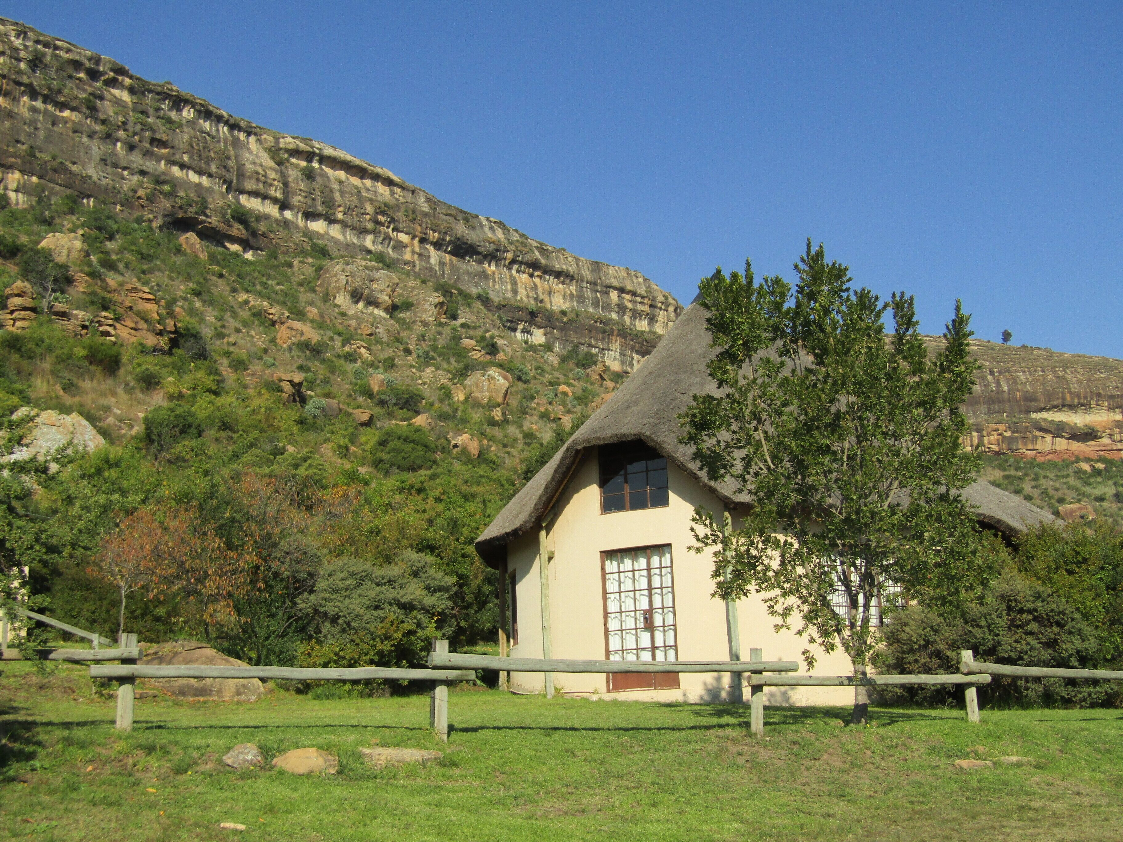 A house with a thatched roof, surrounded by a wooden fence, set against a backdrop of rocky cliffs and greenery.