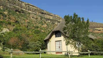 A house with a thatched roof, surrounded by a wooden fence, set against a backdrop of rocky cliffs and greenery.