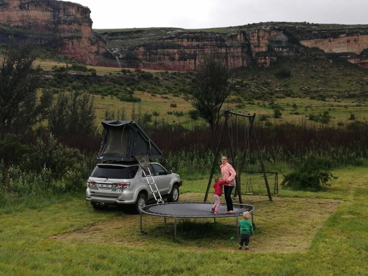 Un coche con una tienda de campaña en el techo, un trampolín y un columpio en un campo de césped con un acantilado al fondo.