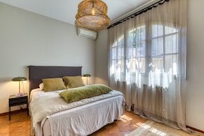 Ein Schlafzimmer mit Bett, Nachttisch, Lampe, einem Fenster mit durchsichtigen Vorhängen und einer Deckenleuchte.