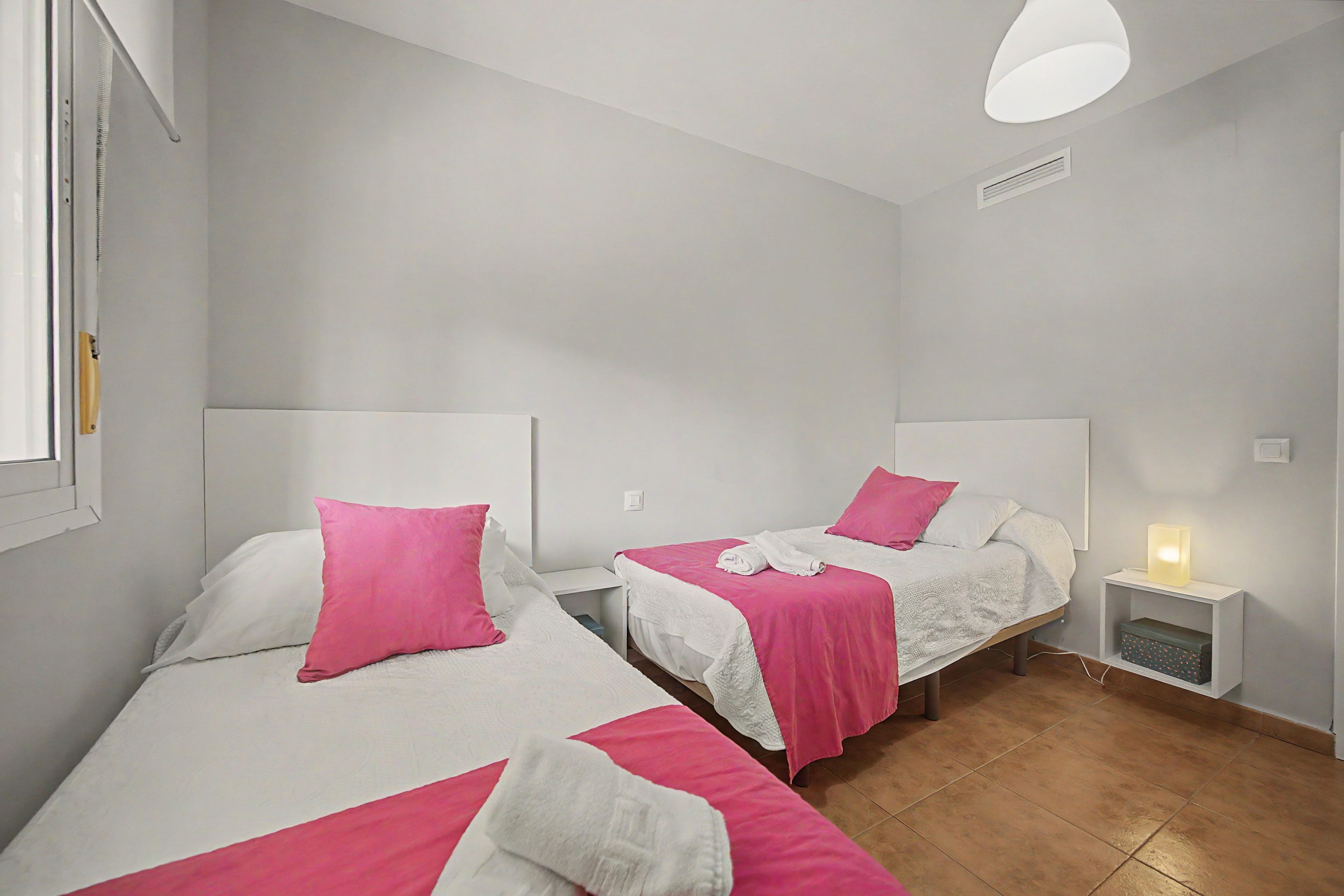 Ein Hotelzimmer mit zwei Betten, jeweils mit einer weißen und rosafarbenen Bettdecke, einem Nachttisch mit Lampe und einem Fenster mit Jalousien.