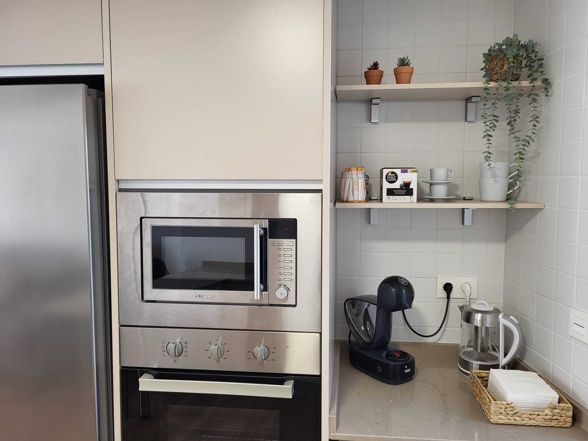 Una cocina con microondas integrado, cafetera y estantes con plantas en macetas y utensilios de cocina.