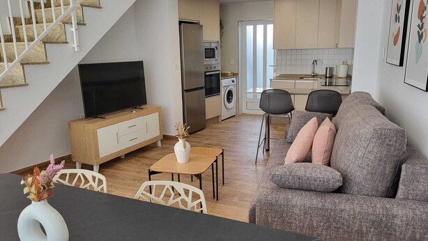 Ett modernt vardagsrum med en soffa, en tv och en matplats. Ett kök med vitvaror syns i bakgrunden.