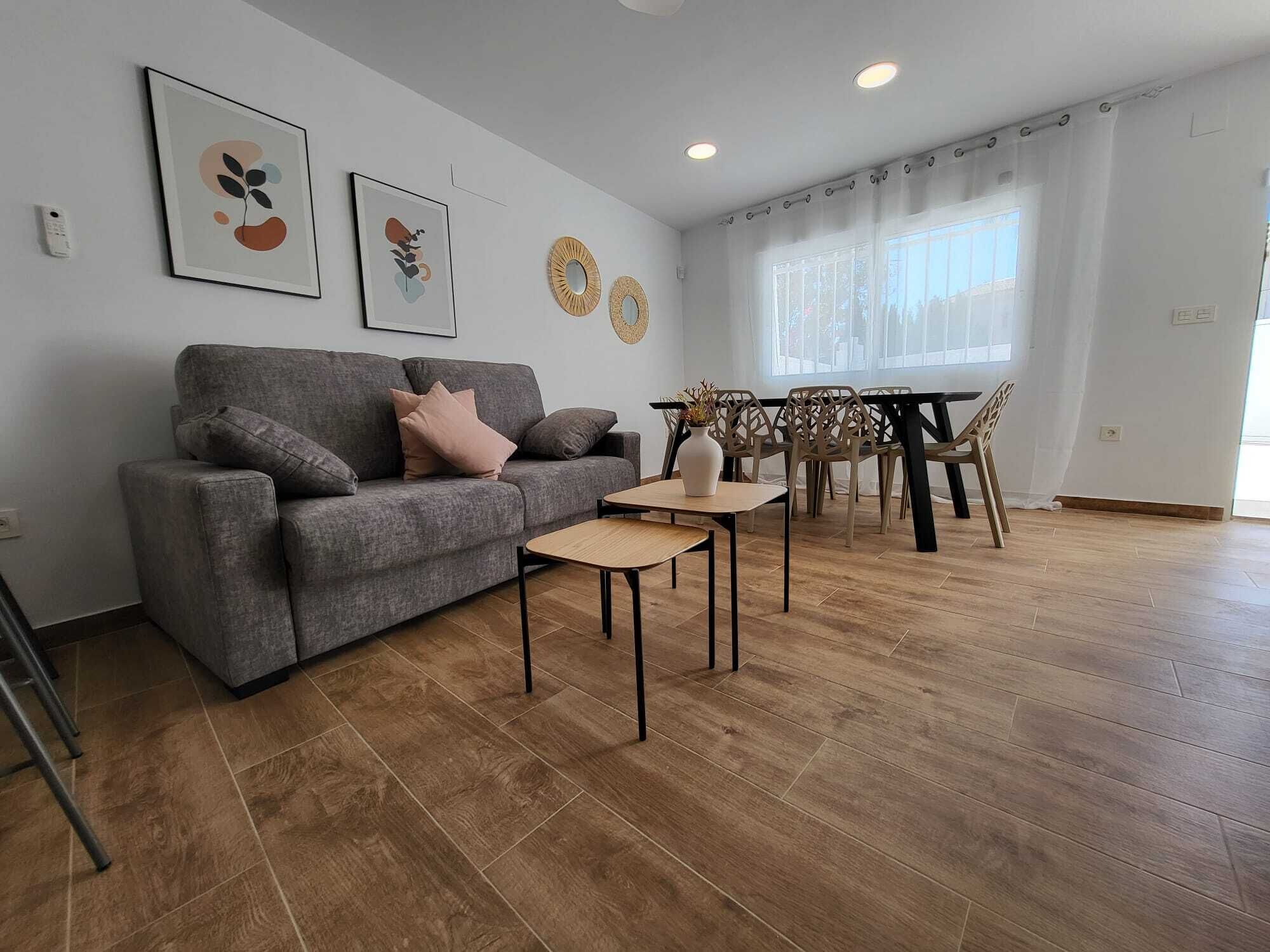 A living room with a grey sofa, a small table, and a dining area with chairs.
