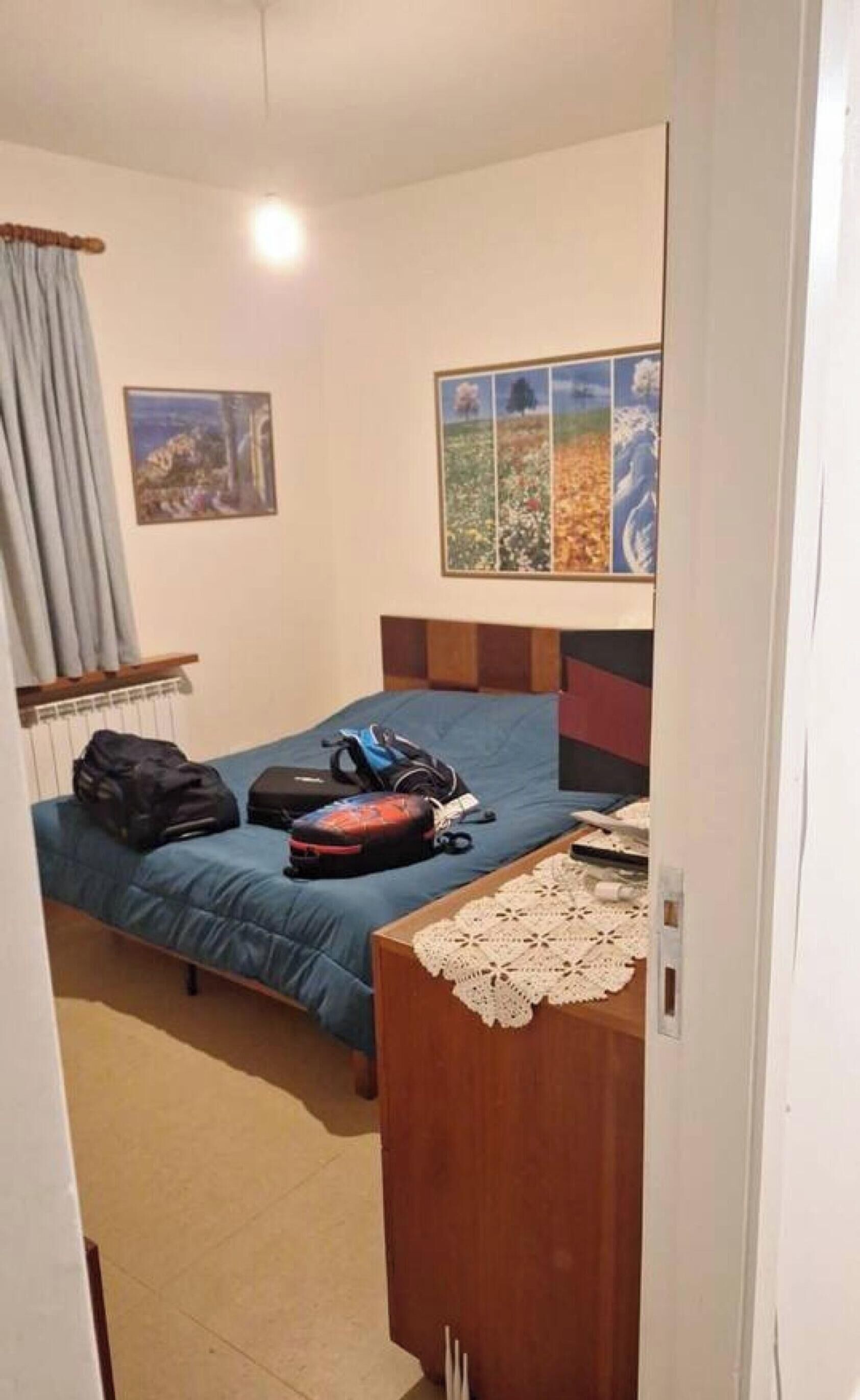 Um quarto com cama, cómoda de madeira, radiador, janela com cortinas e uma parede com pintura de paisagem.