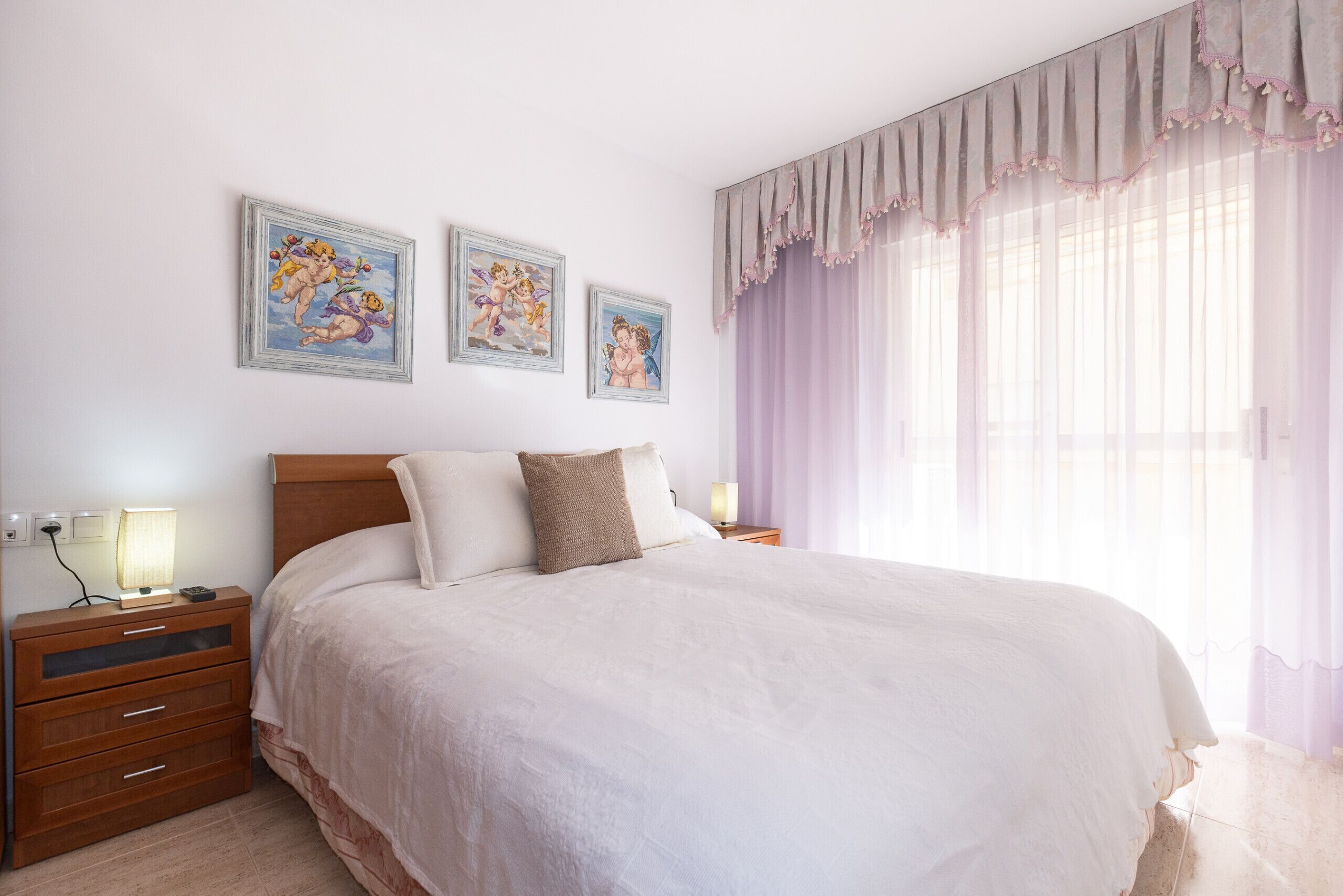 A neatly made bed with a headboard, a bedside table with a lamp, and three framed pictures on the wall.