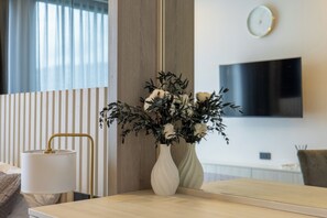 Ein modernes Hotelzimmer mit Bett, Nachttisch mit Lampe, großem Spiegel, Wand-Fernseher und Blumen in einer Vase.