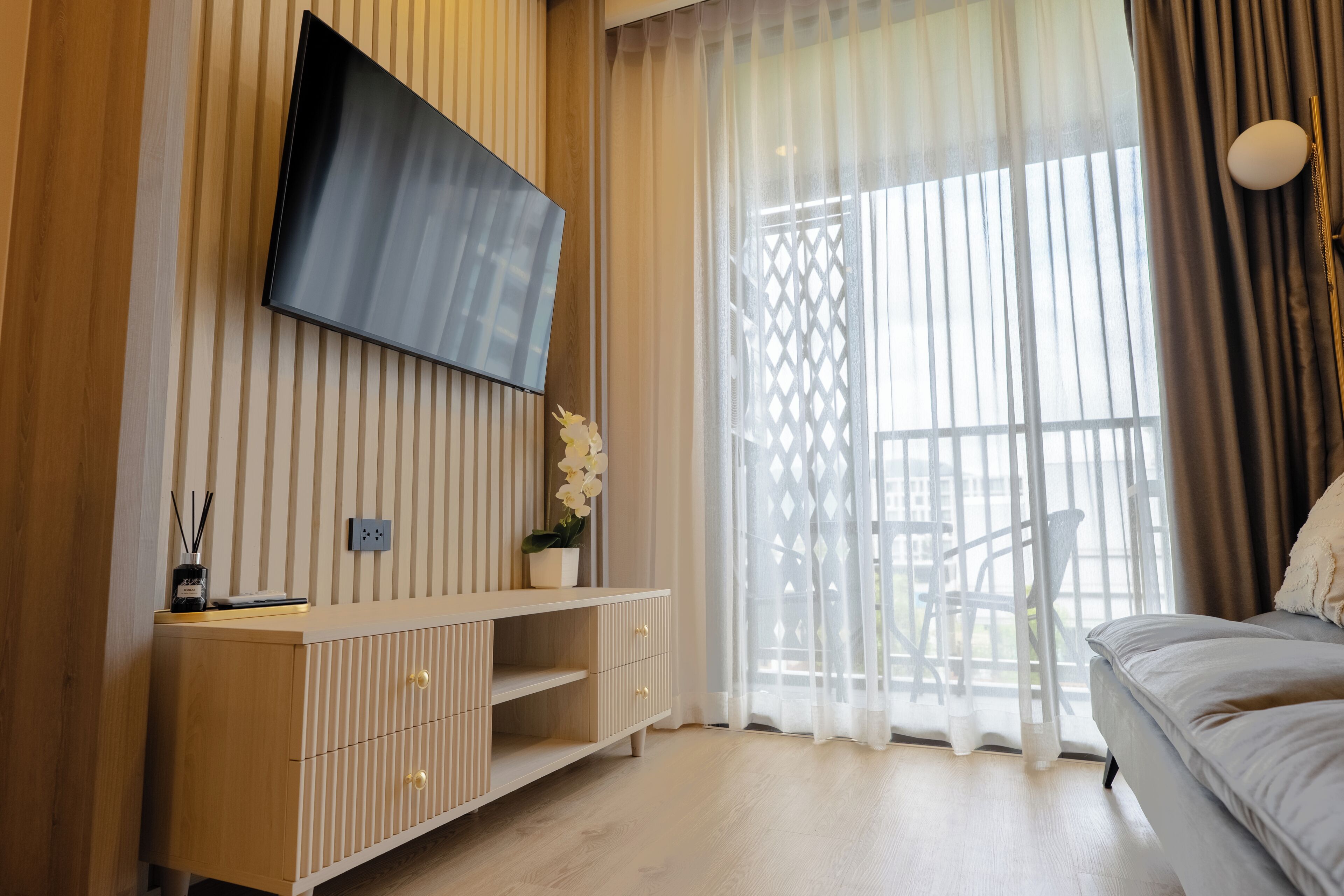 A modern room with a TV mounted on the wall, a wooden cabinet, a bed, and a window with sheer curtains.