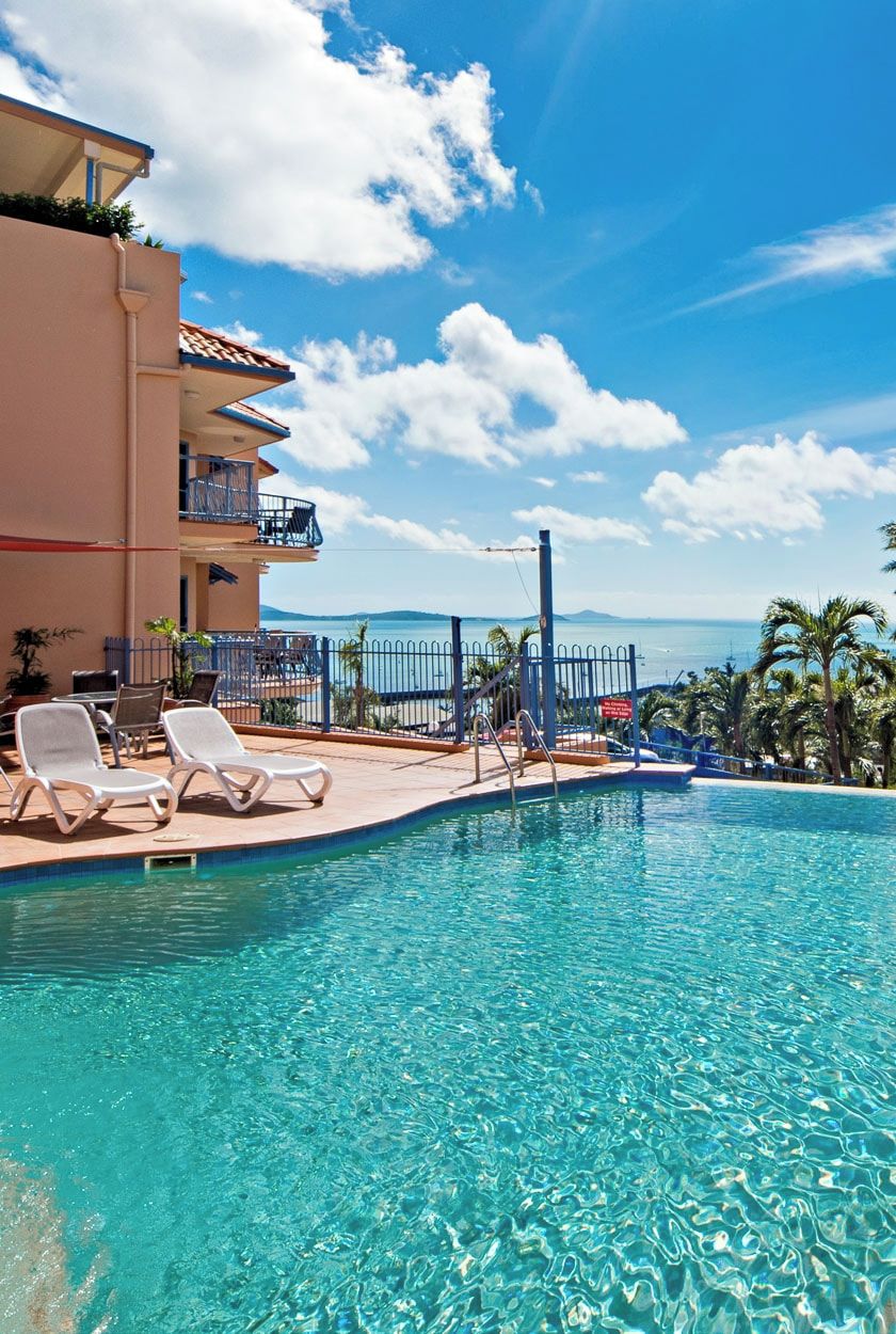 Une piscine d’un bleu éclatant, dotée de chaises longues, un balcon avec vue sur la mer et un ciel dégagé.