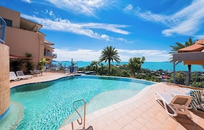 Ein Swimmingpool mit Liegestühlen, Blick auf das Meer und ein Gebäude mit Balkonen.