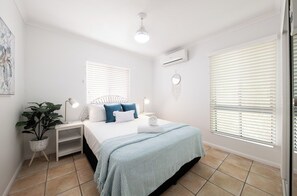 A bedroom with a bed, bedside tables, a lamp, a potted plant, and a wall-mounted air conditioning unit. - Sea Crest (Airlie Beach)
