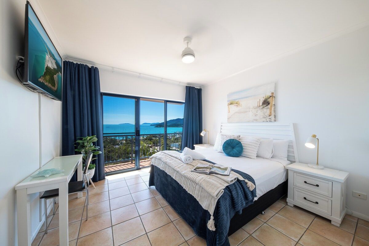 A bedroom with a bed, bedside tables, a desk, and a large window offering a view.